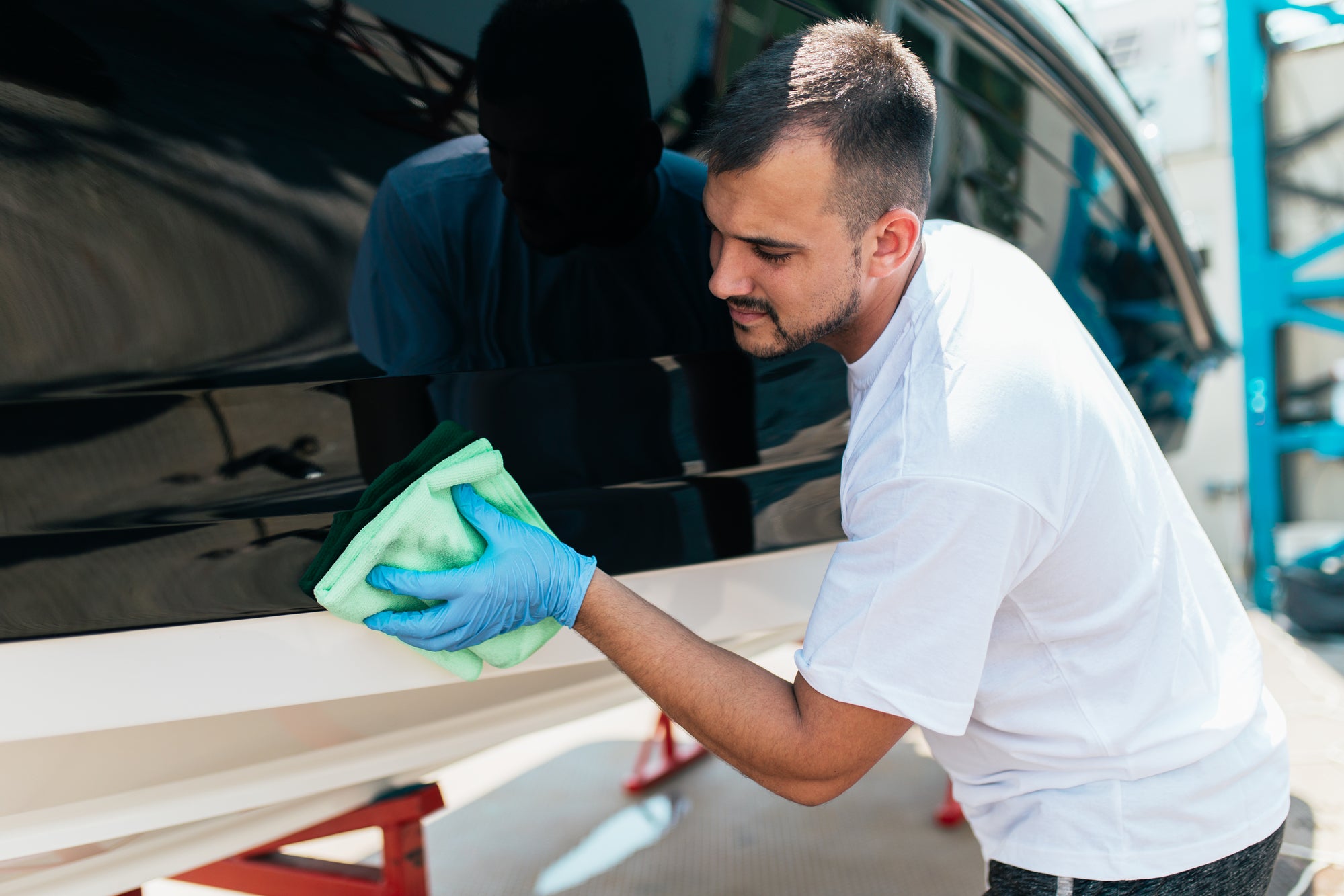 Man cleaning a black vehicle with a green microfiber cloth, wearing blue gloves, in a professional detailing setting.