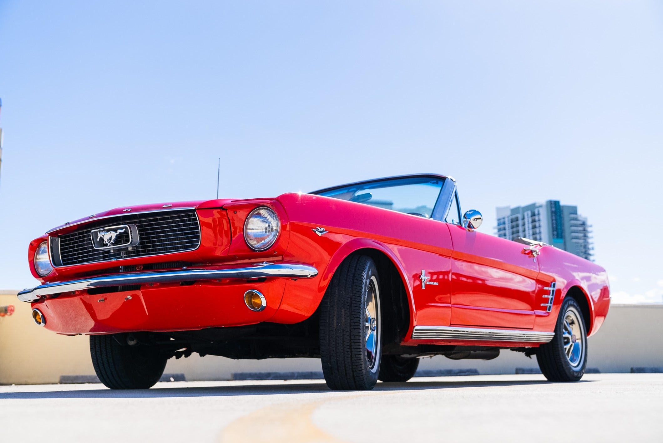 Red vintage Ford Mustang convertible parked outdoors with a clear blue sky and city buildings in the background.