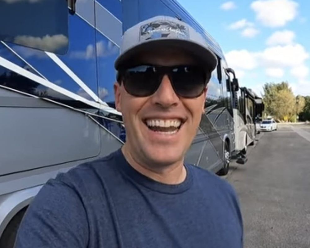 Smiling man in sunglasses and cap outside a black and silver luxury RV, parked on a sunny day with a clear sky.