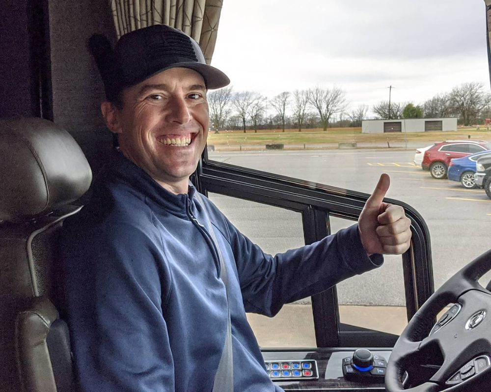 Smiling man in a blue jacket giving a thumbs-up inside a vehicle, with a parking lot and overcast sky outside.