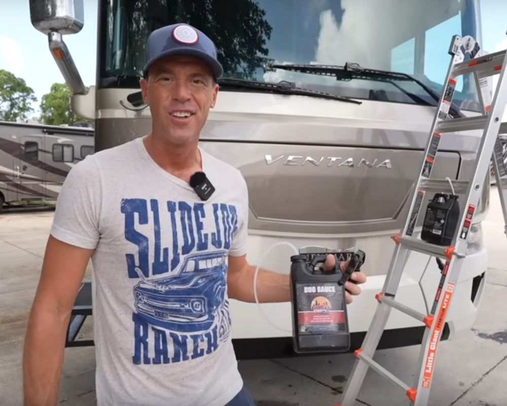Man holding vehicle cleaning products in front of a motorhome, with ladder and equipment for professional car detailing.