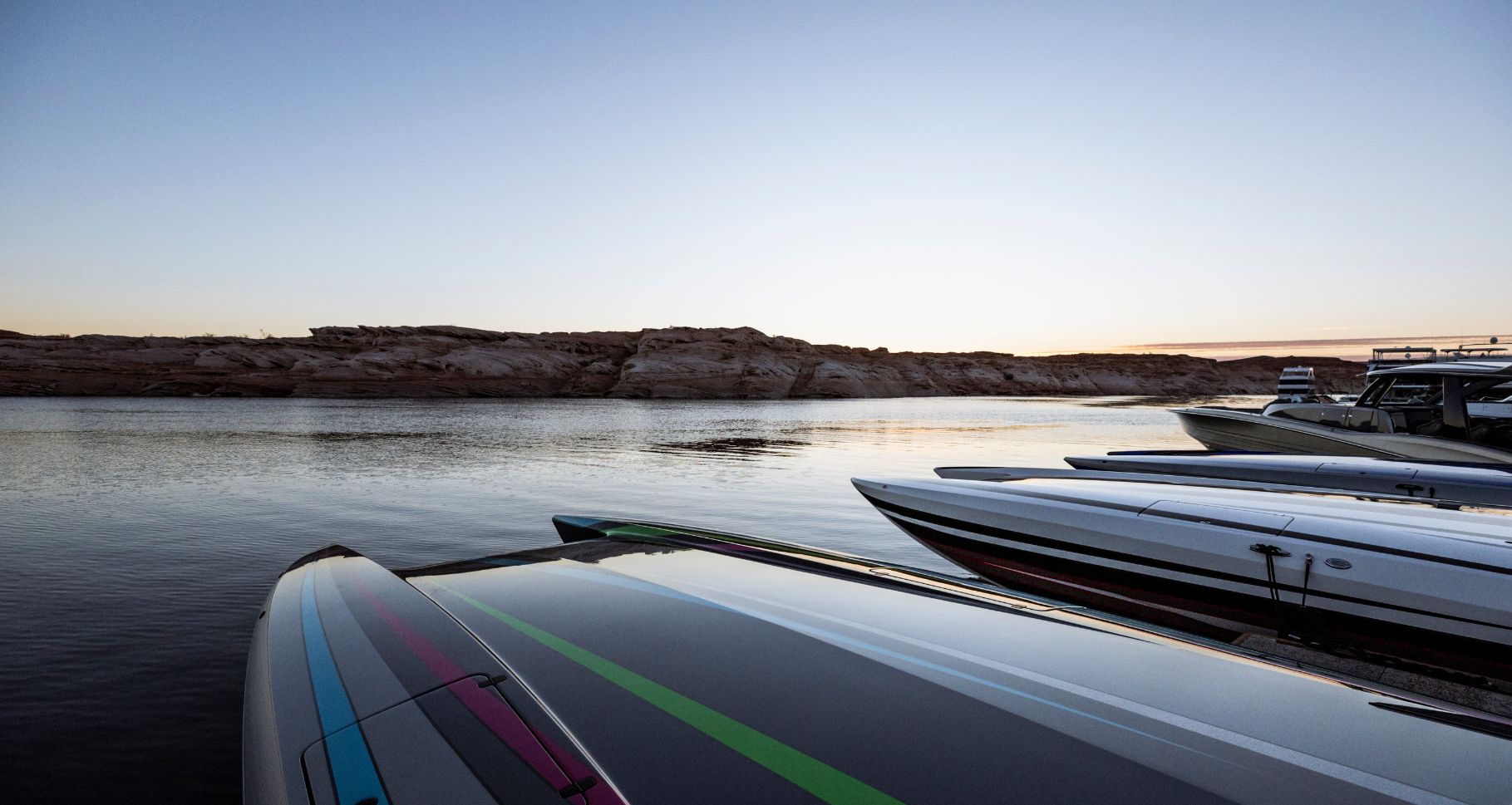 Multiple sleek boats docked on calm water at sunset, with rocky shoreline in the background, showcasing premium marine veh...