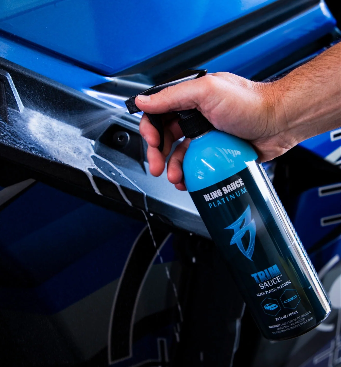 Hand spraying blue "Trim Sauce Platinum" plastic restorer on a vehicle's black trim.