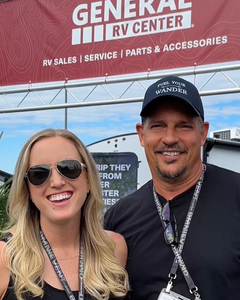 Smiling man and woman at an RV center event, woman wears sunglasses, man has a "Fuel Your Wander" cap, both with event lan...