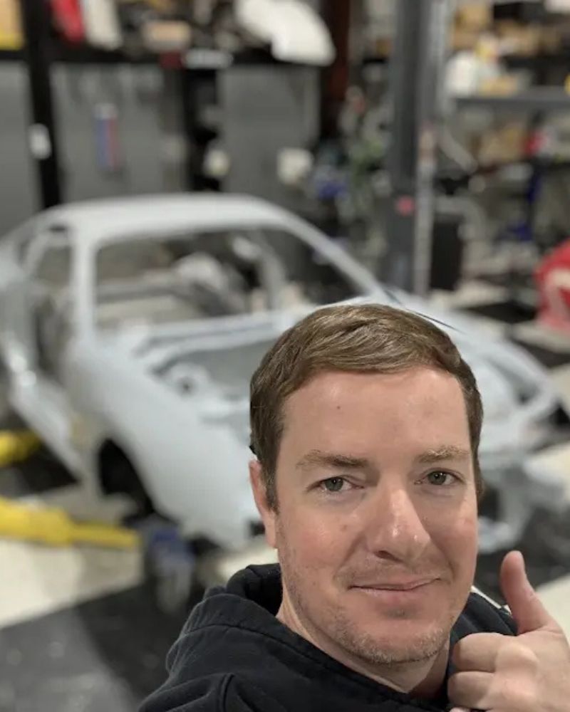 Man giving a thumbs-up in front of a white, partially assembled sports car in a garage or workshop.