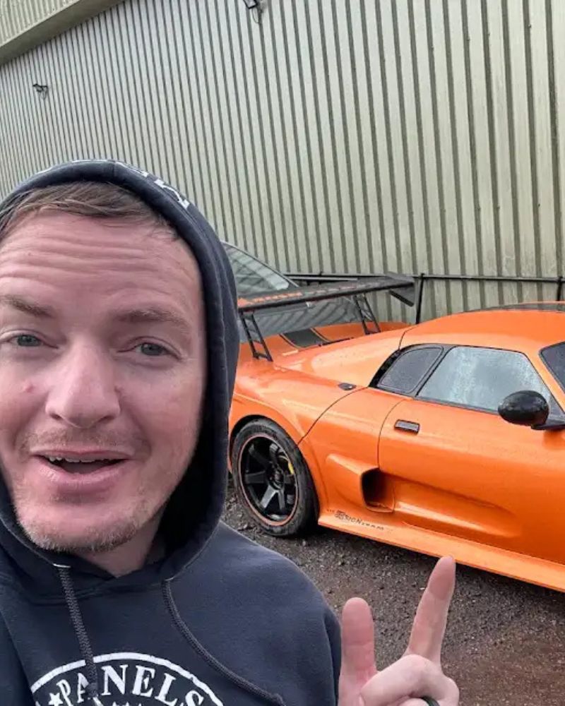 Close-up of a man in a hoodie pointing at an orange vintage race car, parked outside next to a corrugated metal wall.