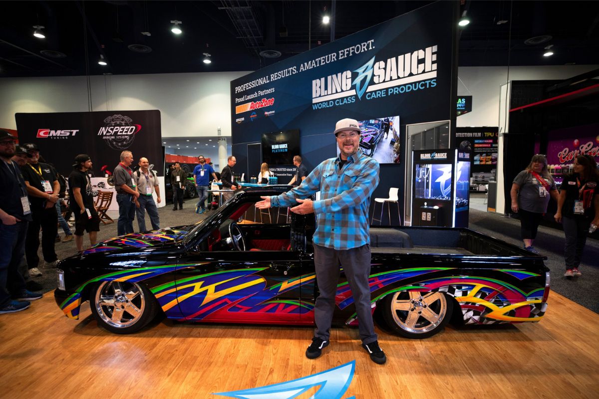 Man in plaid shirt showcasing a custom black pickup truck with colorful, vibrant graphic wrap at a vehicle detailing expo.