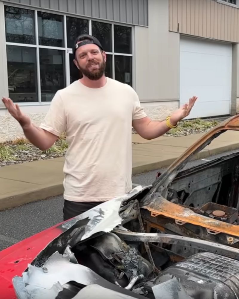 Man smiling with arms outstretched next to a burnt, damaged vehicle in front of a modern building, promoting premium vehic...