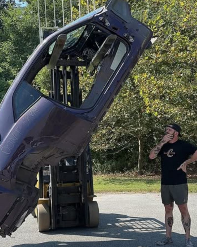 A man in black shorts and a cap stands next to a forklift with its cab tilted forward, surrounded by greenery.