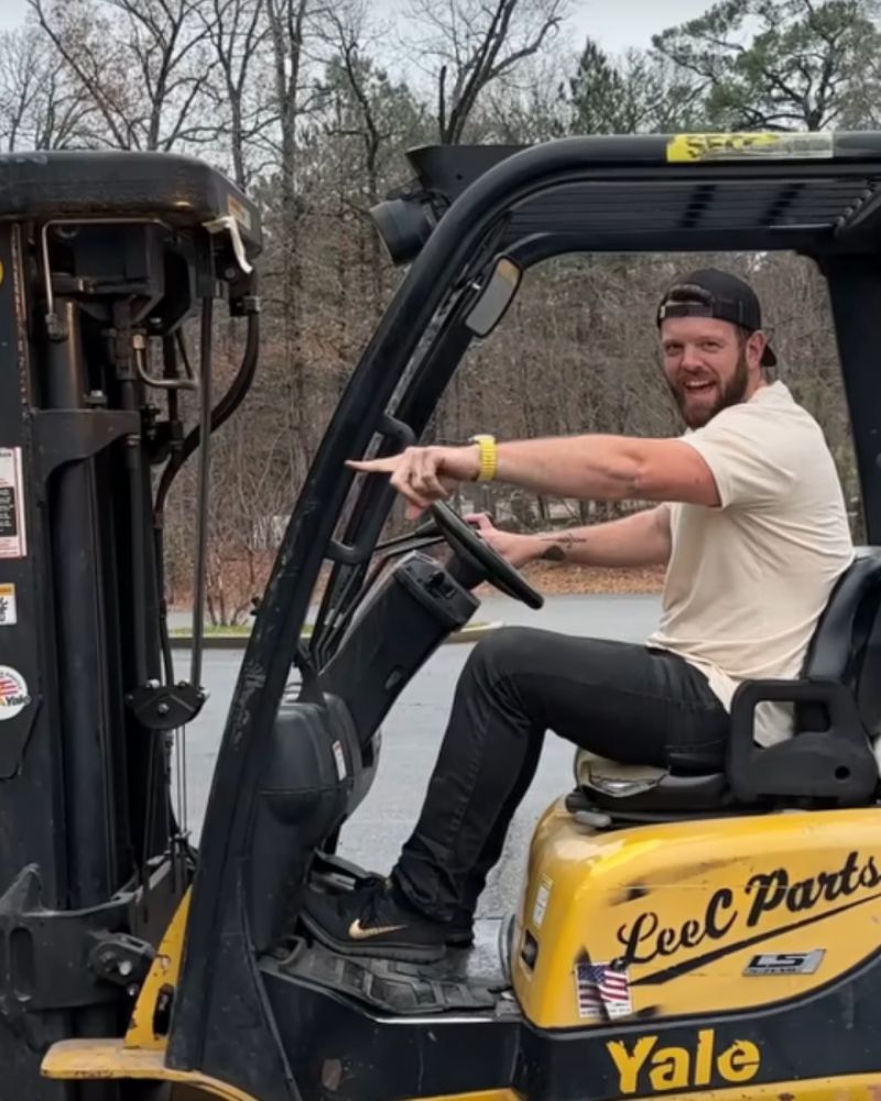 Happy man in a white shirt and black cap riding a yellow Yale forklift, pointing and smiling outdoors.