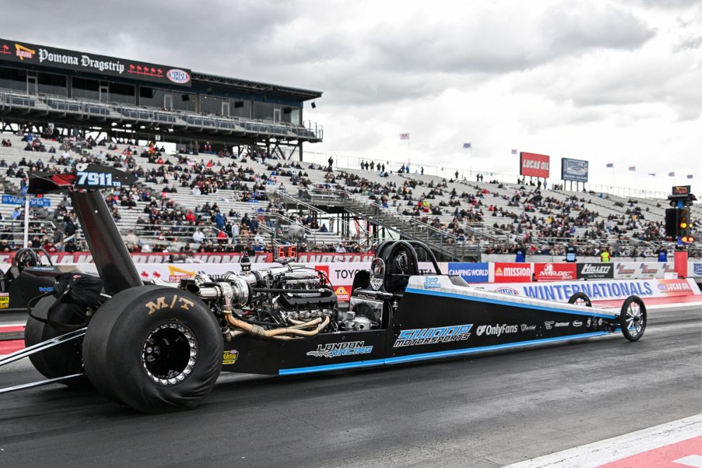 High-speed dragster on track at a race, with black and blue body, large rear tires, and racing decals, under cloudy skies.