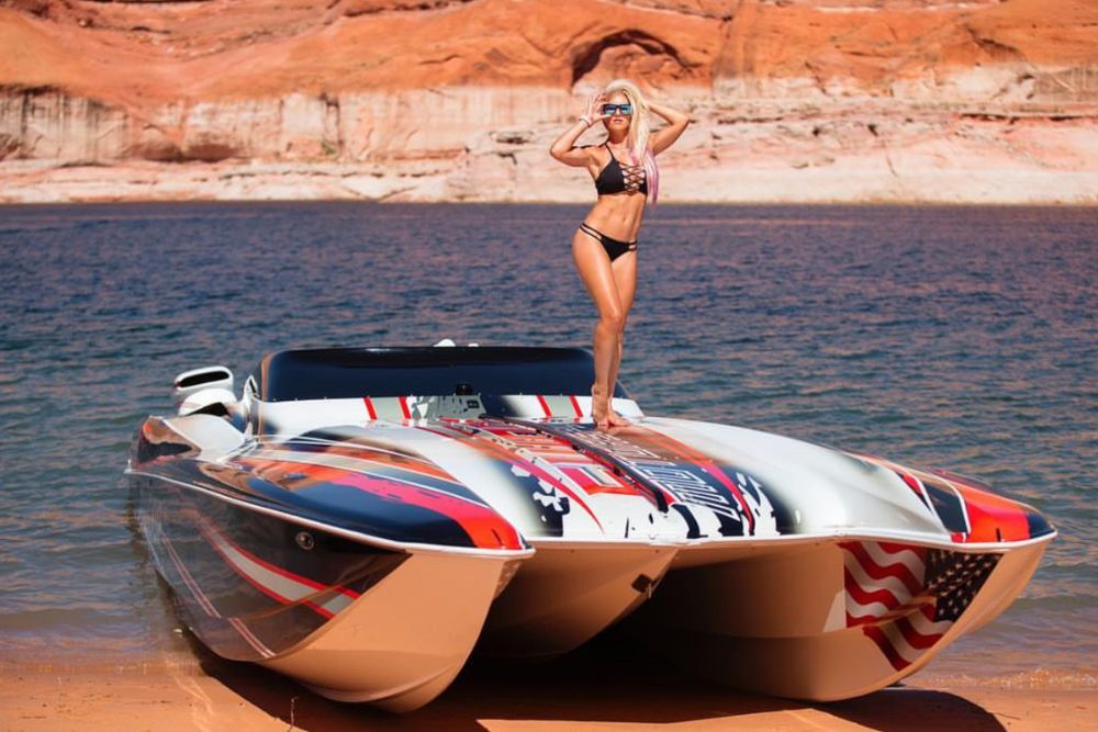 Woman in black swimsuit on a patriotic speedboat, with red, white, and blue accents, parked on a sandy beach near water.