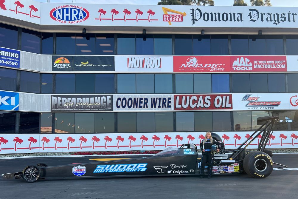 Drag racing car with sponsor decals, black and blue color, parked in front of a building with multiple automotive and raci...