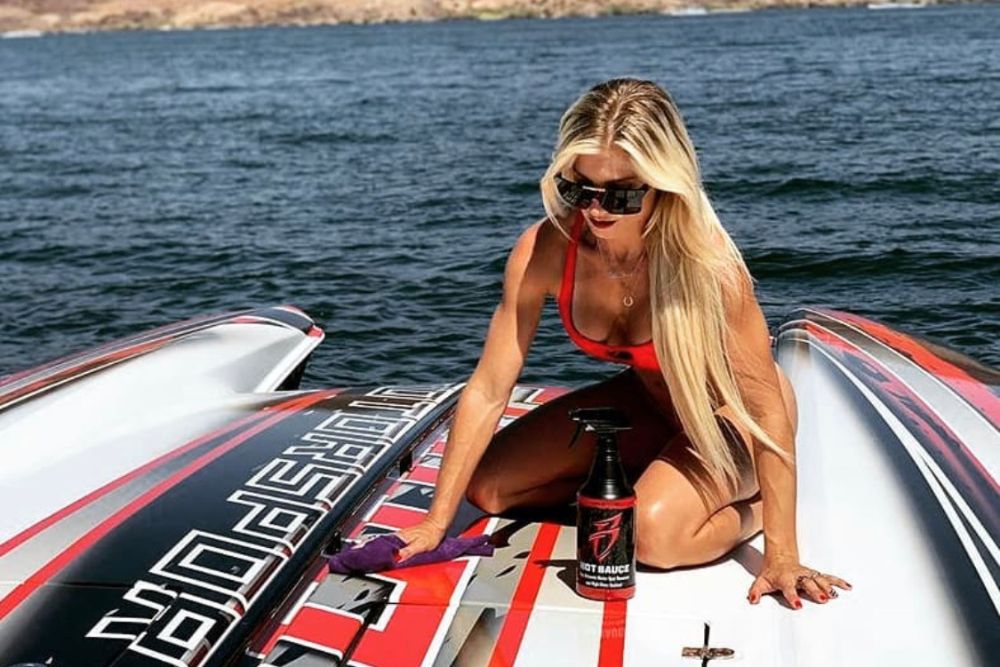 Woman cleaning a boat with a black and red spray bottle, wearing sunglasses and a red top, on a boat with a colorful design.