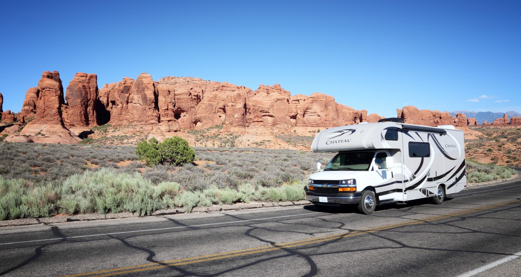 White RV with black and gray detailing parked on a desert road with red rock formations and clear blue sky.