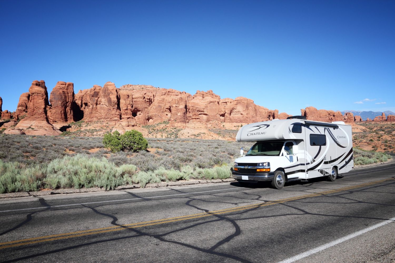 White RV with black and gray swoosh decals parked on a desert highway with red rock formations and blue sky.