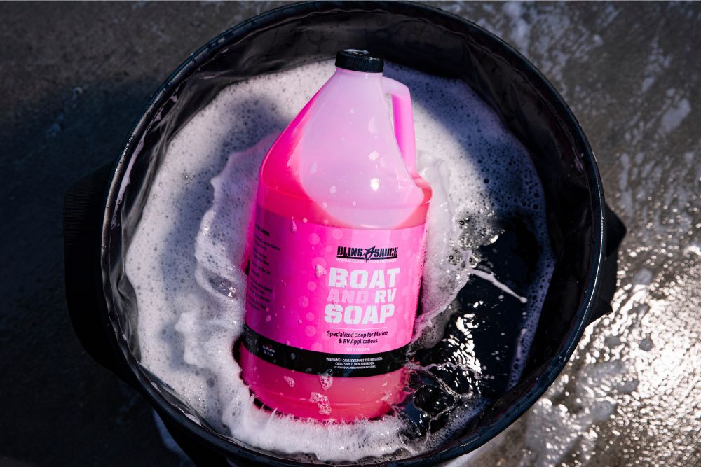 Bottle of Bling Sauce Boat and RV Soap surrounded by suds in a bucket, showcasing its vibrant pink color and label.