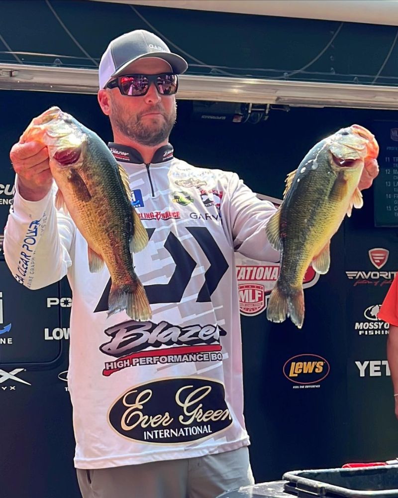 Man holding two large bass fish, wearing sunglasses and a branded fishing jersey, against a backdrop of fishing sponsors.