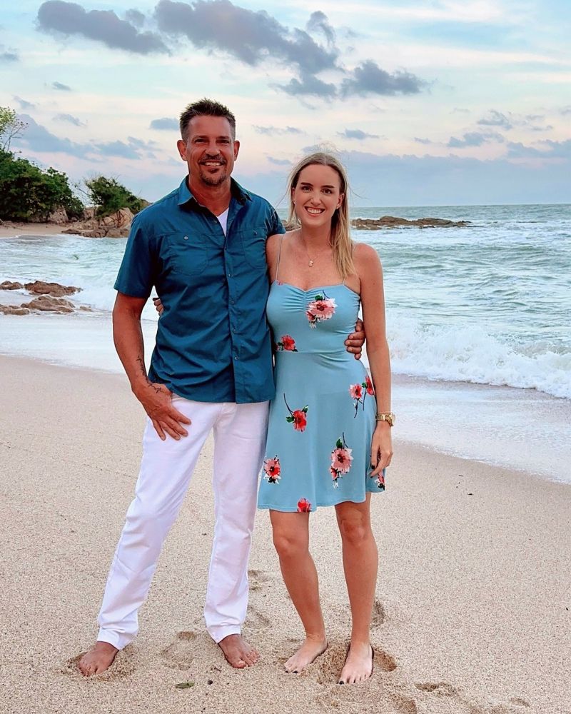 Two smiling people standing barefoot on the beach, with ocean and cloudy sky in the background. Woman in a blue floral dre...