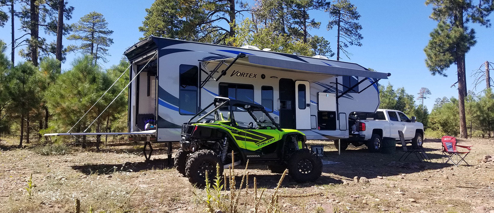 Bright green off-road ATV parked in front of a large blue and white RV trailer in a wooded outdoor setting.