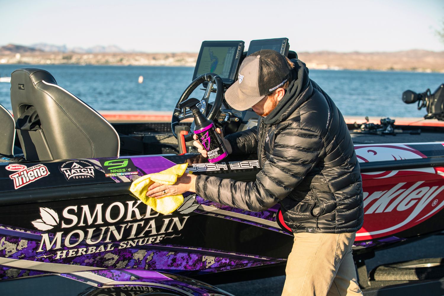 Man cleaning a boat with a yellow microfiber towel and purple vehicle detailing spray, near a lake, promoting premium vehi...