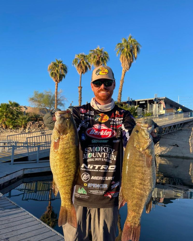 Man holding two large bass fish, wearing a branded fishing jersey and cap, with palm trees and a dock in the background.