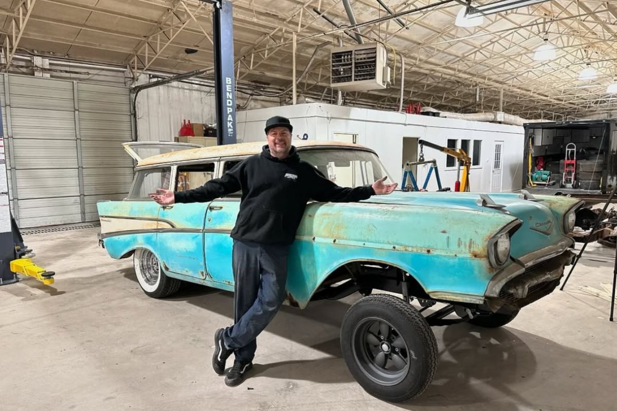 Man in black hoodie posing with a vintage, rusty turquoise car inside a workshop, emphasizing vehicle restoration and deta...