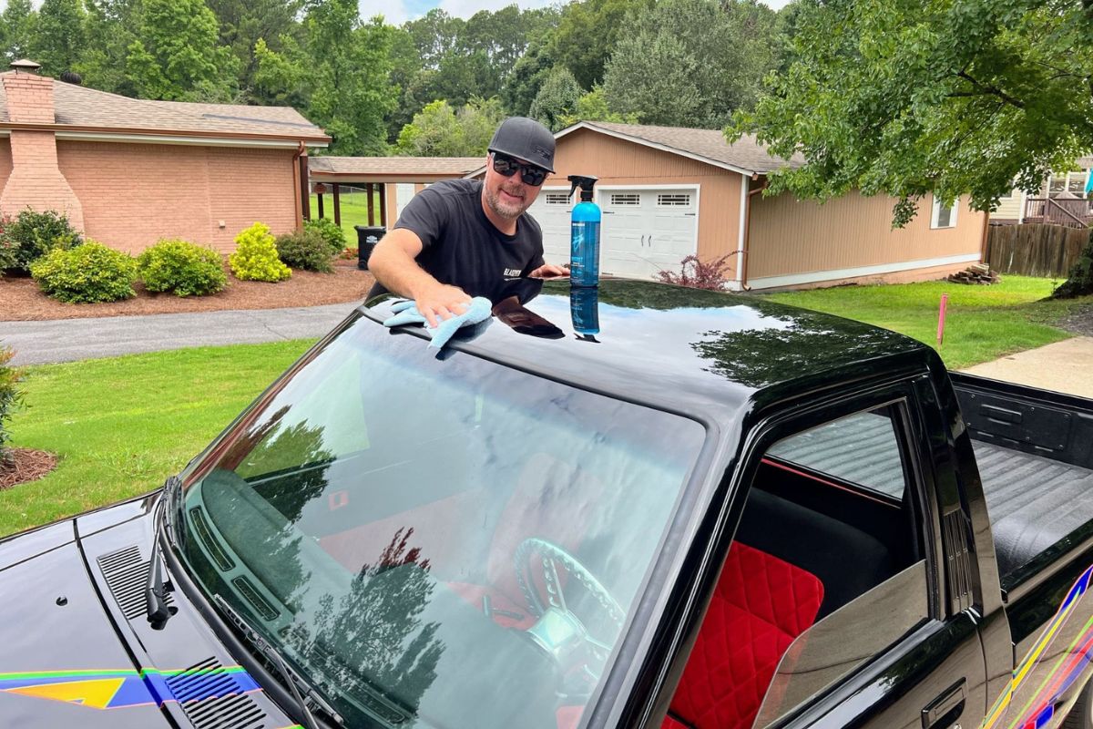 Man cleaning black vehicle windshield with microfiber cloth, blue detailing spray bottle on roof, in a suburban driveway.