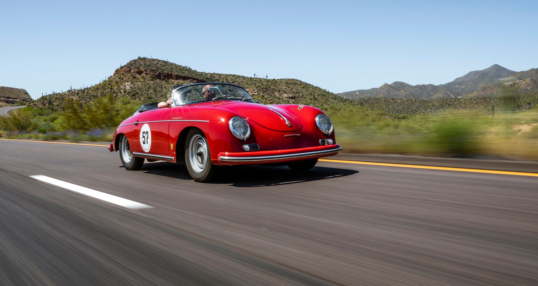 Red vintage Porsche sports car driving on open road with desert mountains in background, emphasizing high-performance vehi...