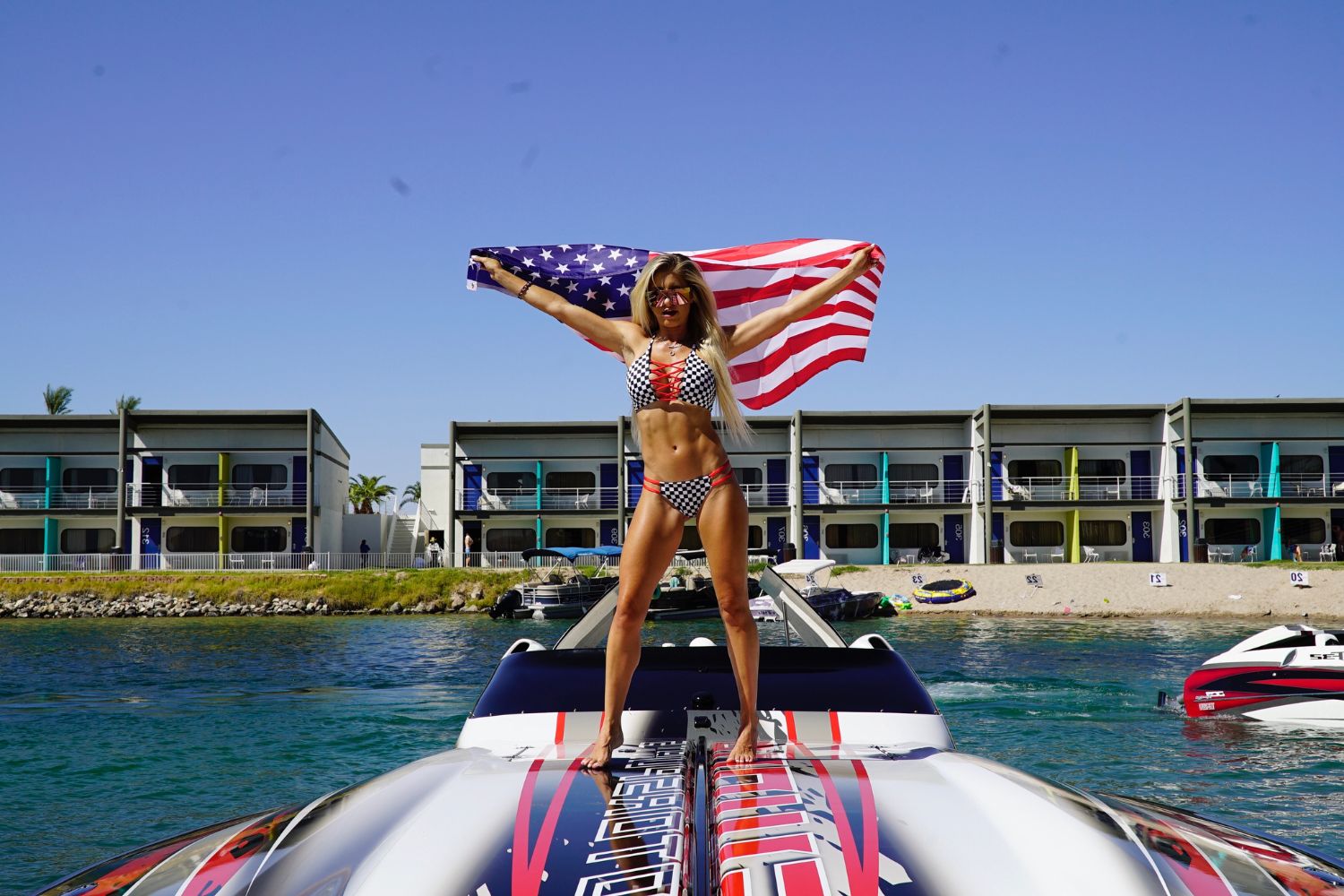 Woman in checkered bikini holding American flag on a boat, with colorful modern buildings and clear blue sky in the backgr...
